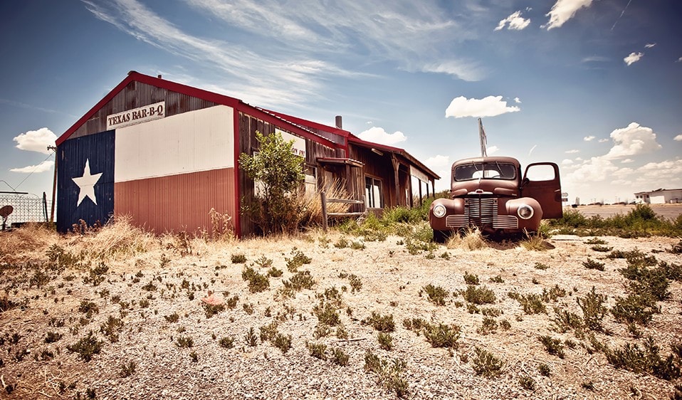 An old truck sits in front of a Texas flag building.